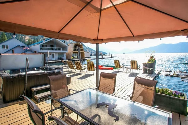 Waterfront deck with orange umbrella, dining table, lounge chairs, and lake view with mountains and boathouses