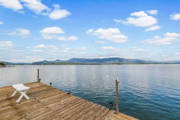 Wooden dock with white Adirondack chair extending into calm lake, mountains visible across water under blue sky with white clouds